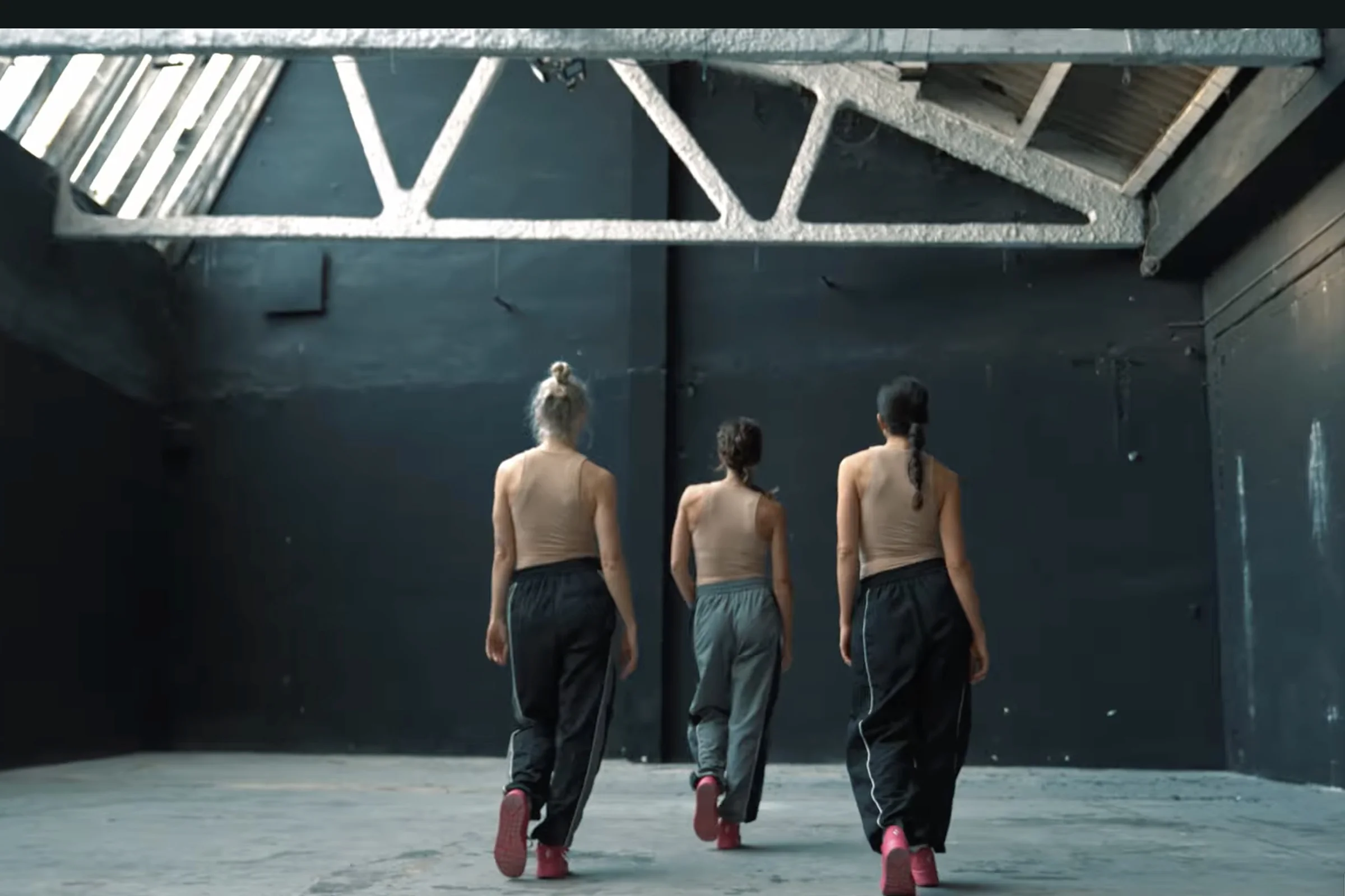Tres mujeres de espaldas hacen el mismo gesto, con el pie izquierdo delante del derecho. Llevan tops sin mangas de color carne y pantalón deportivo.