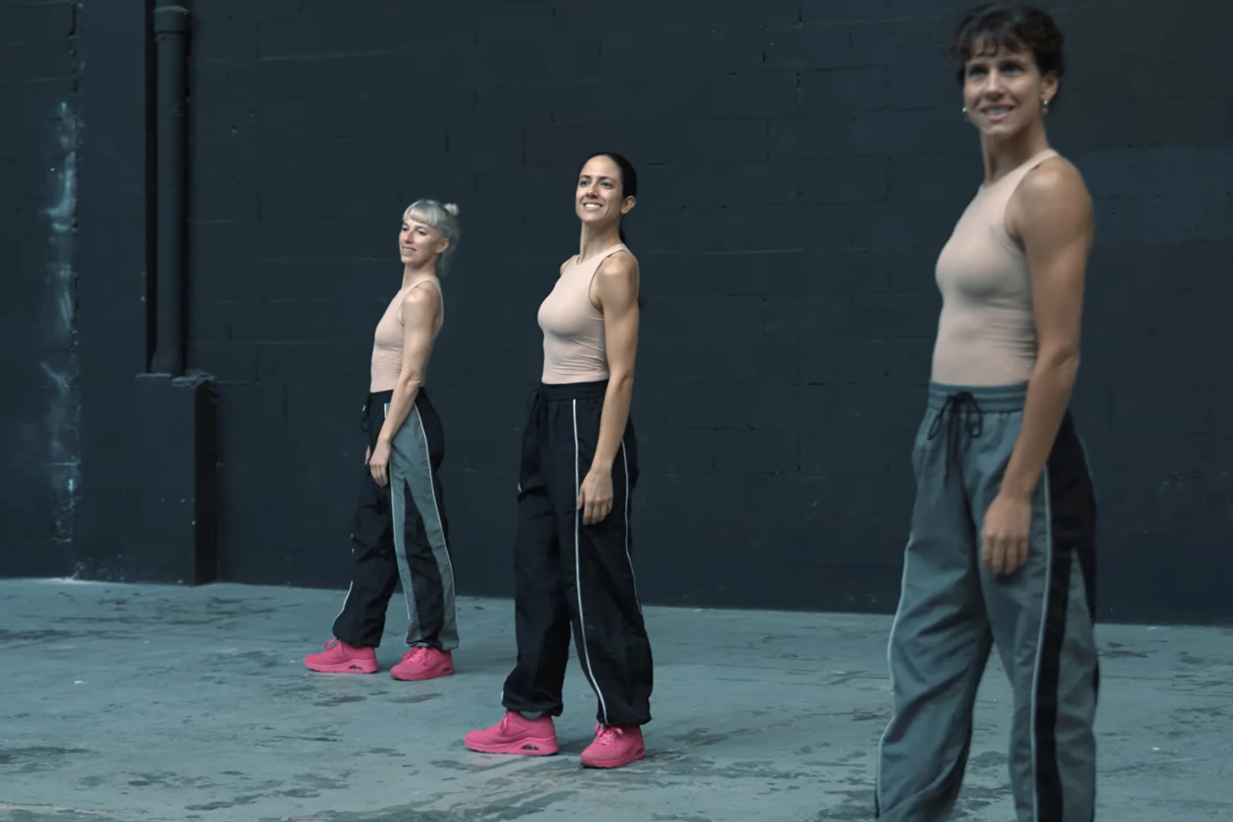 Tres mujeres con una sonrisa forzada. Las tres están de pie y llevan tops sin mangas de color carne y pantalón deportivo.