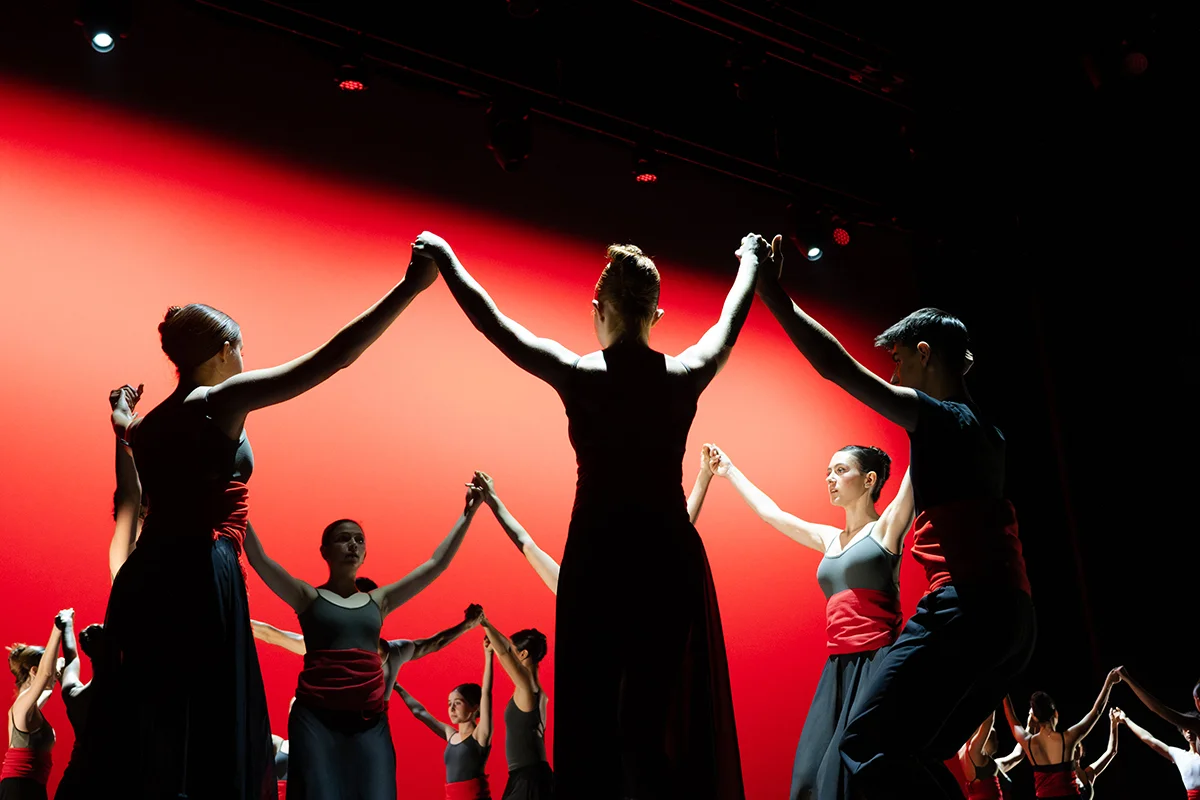 Tres corros de jóvenes bailan una sardana sobre el escenario de un teatro. El fondo es rojo.