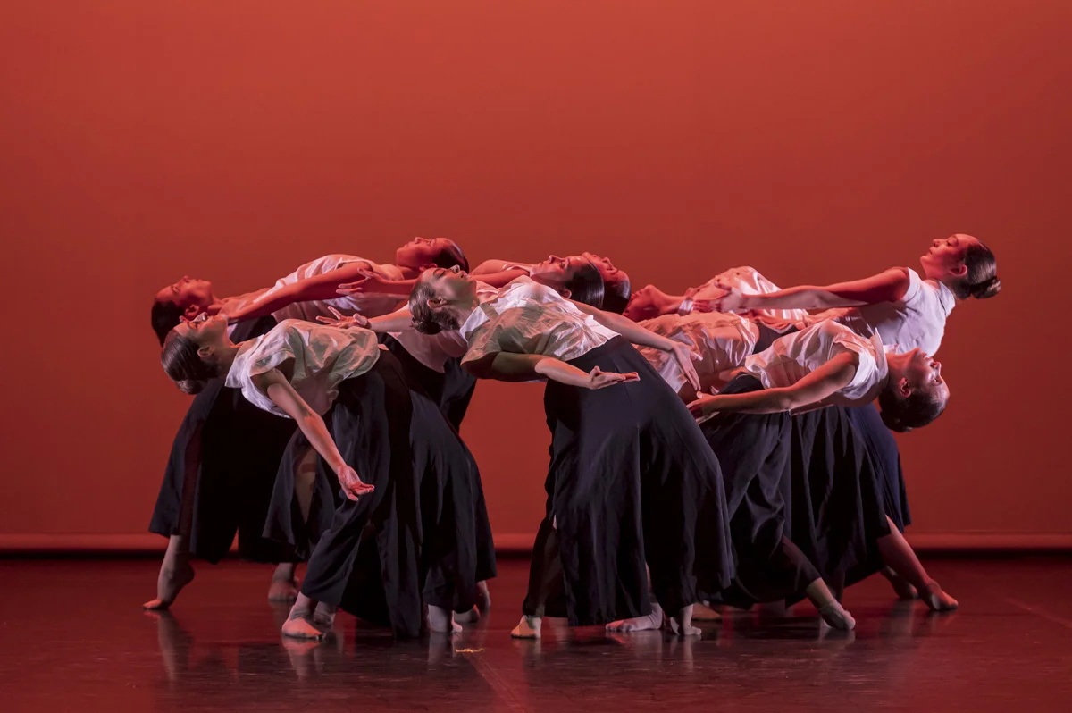 Siete chicas realizan una coreografía sobre fondo rojo. Están en círculo y echan la cabeza hacia atrás todo lo que pueden.