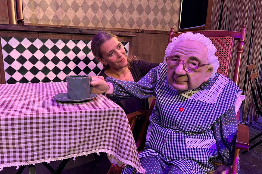 Un titella d'una àvia seu i pren un cafè. També apareix a la fotografia l'actriu que manipula el titella.