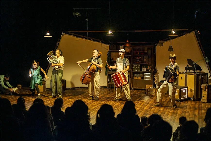Sobre l'escenari, quatre músics toquen tuba, violoncel, tambor i acordió, mentre una cinquena persona corre amb una bombeta a la mà.