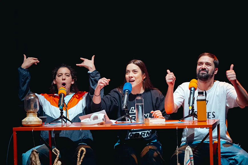 Dues noies i un noi seuen a una taula davant d’uns micròfons en format pòdcast.