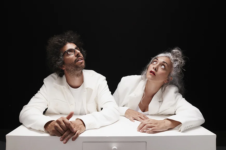 Escena de l’espectacle Nozing, on es veuen els dos integrants del dúo Ual·la! asseguts i mirant cap amunt davant d’una taula blanca.