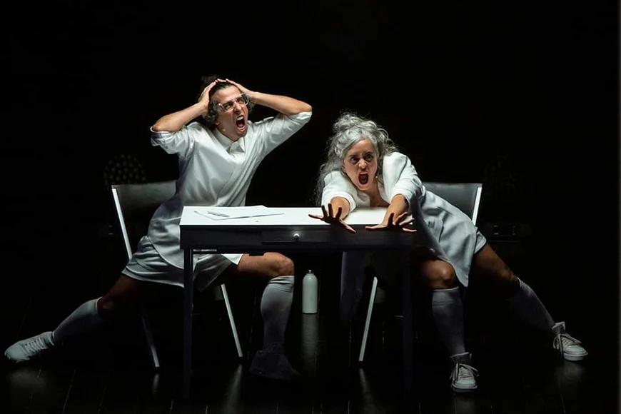 Escena de l’espectacle Nozing, on es veuen els dos integrants del dúo Ual·la! amb cara de por davant d’una taula blanca.