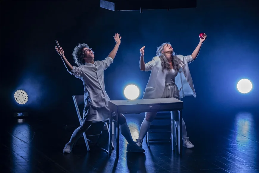 Escena de l'espectacle Nozing, on es veuen els dos integrants del dúo Ual·la! dempeus i amb els braços aixecats devant d'una taula blanca.