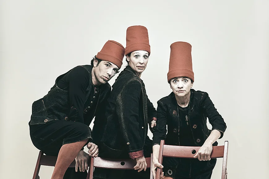 Tres personatges vestits de negre i amb gorros vermells alts seuen en cadires plegables i miren a càmera sobre un fons blanc.