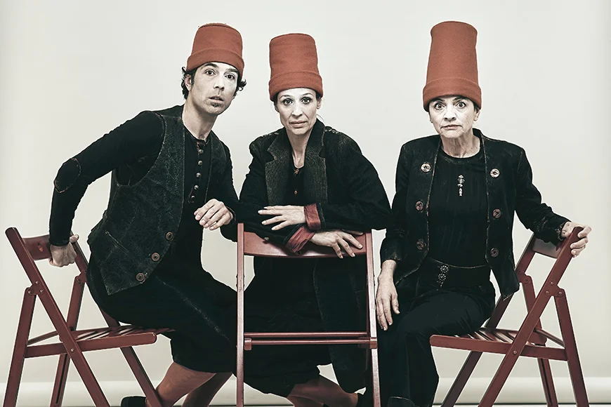 Tres personatges vestits de negre i amb gorros vermells alts seuen en cadires plegables i miren a càmera sobre un fons blanc.