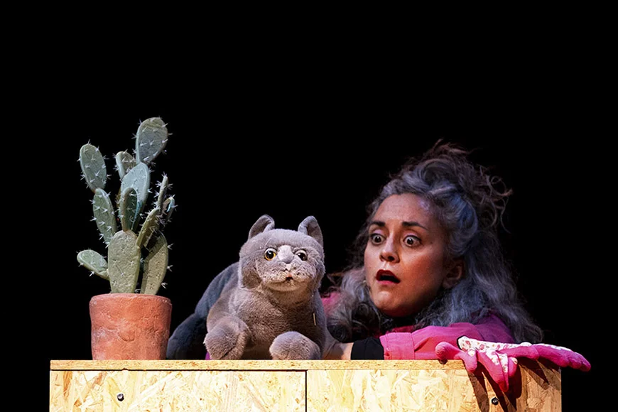 La protagonista de l'espectacle mira amb cara de sorpresa el gat de peluix que manipula. A la imatge també hi veiem un cactus.